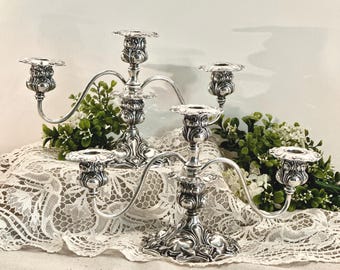 May include: Two silver-colored metal candelabras, each featuring three candle holders with curved arms and ornate, embossed floral detailing on the base and stems. The polished metallic finish reflects light, highlighting the intricate scrollwork.