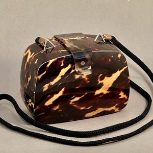 May include: A box-shaped handbag featuring a glossy brown and cream tortoiseshell pattern. The bag has a hinged top closure with a matching tortoiseshell tab and a long, thin black cord strap attached with silver-toned metal hardware.