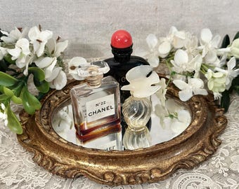 May include: Three glass perfume bottles on a gold-toned oval mirror tray. One bottle is square with a label reading N°22 CHANEL, one is black with a red cap, and one is small and clear with a bird-shaped stopper, surrounded by white flowers.