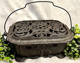 May include: A cast iron trivet with an ornate, openwork design on the top and decorative detailing on the sides. It has a rectangular shape with a handle and is dark gray in color.