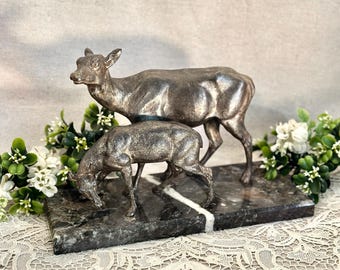 May include: A metallic silver-toned sculpture of two deer standing on a rectangular dark gray marble base with a prominent white vein running through the stone. The larger deer stands upright while the smaller deer is positioned in front, grazing.