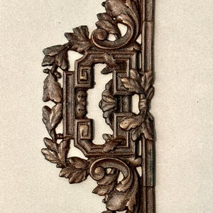 May include: A decorative metal ornament with a detailed design of leaves and scrolls. The ornament is in a dark brown color and has a geometric pattern in the center.