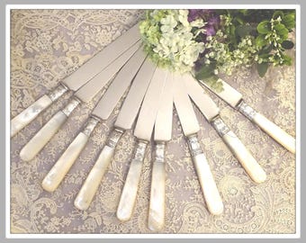 May include: A set of eight silver-toned knives arranged in a fan pattern. Each knife features a smooth, polished metal blade and a handle crafted from iridescent white mother-of-pearl with an ornate silver-colored metal collar at the base.