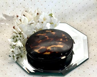 May include: A round compact case featuring a glossy dark brown and amber tortoiseshell pattern with a smooth finish. The compact is positioned on a reflective octagonal mirror surface, accompanied by a cluster of white artificial flowers.
