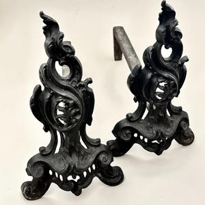 May include: Pair of antique black cast iron fireplace andirons with ornate, scrolled designs. The andirons have a decorative base and a curved bar to hold logs. The andirons are a decorative and functional addition to a fireplace.
