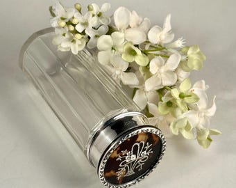 May include: A clear glass bottle with vertical fluting and a silver-toned metal cap. The cap features a circular tortoiseshell-patterned inlay with a white engraved floral basket design and a decorative silver border, accompanied by white flowers.