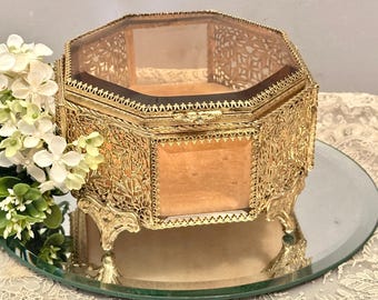 May include: An octagonal jewelry box featuring a gold-tone metal frame with intricate filigree detailing and beveled glass panels. The box is supported by four ornate feet and includes a hinged lid with a latch, revealing a soft, light-colored interior.
