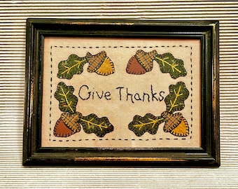 May include: A rectangular framed wall art piece featuring a light beige fabric background with the text Give Thanks embroidered in black. The design includes four corners decorated with appliqued green oak leaves and brown and yellow acorns.