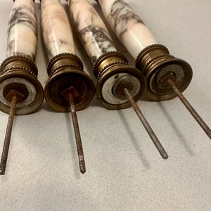 K&ouml;nnte beinhalten: Vier antike Schubladengriffe aus Marmor und Messing. Der Marmor ist wei&szlig; mit grauen und rosa Adern. Das Messing hat einen dekorativen Rand und eine Gewindestange, die sich von der Basis erstreckt. Die Griffe sind in einer Reihe angeordnet.