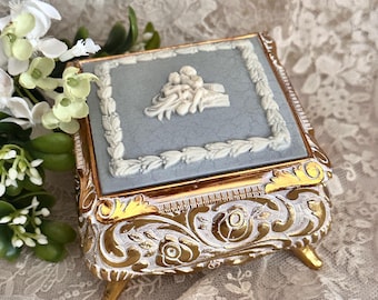 May include: A square jewelry box featuring a light blue lid with a white relief of figures and a floral border. The sides of the box are white with gold-painted ornate floral scrollwork and four small feet. The lid has a visible crackled texture.