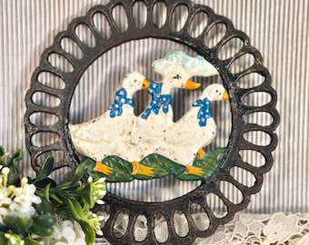 May include: A round, black cast iron trivet featuring a decorative openwork border. The center depicts three white geese with orange feet and beaks, wearing blue and white polka-dot neckerchiefs, walking across a green grassy base.