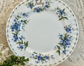 May include: A white ceramic plate with a scalloped edge, decorated with blue forget-me-not flowers and green leaves. The word July is written in gold script at the top center. The plate features a delicate floral pattern around the rim.