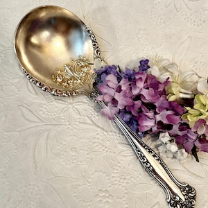 May include: A silver serving spoon with ornate detailing on the handle and bowl edge. The spoon is positioned next to a cluster of purple, white, and yellow flowers, creating a decorative arrangement. The spoon's design is vintage.