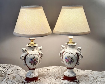 May include: A pair of table lamps featuring white ceramic bases with ornate scrollwork, deep red accents, and hand-painted floral motifs. Each lamp has a gold-toned metal neck and a tapered, light-colored fabric lampshade that emits a soft glow.
