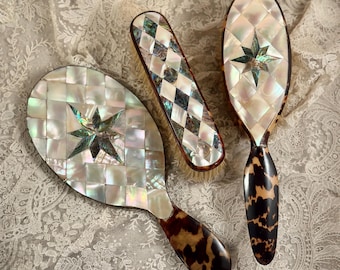 May include: A set of three grooming tools including a hand mirror, a rectangular brush, and an oval hairbrush. Each piece features a mosaic of iridescent mother-of-pearl tiles in a diamond pattern with a central star and tortoise shell handles.