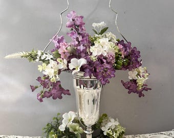 May include: A silver-toned metal pedestal vase with a tall, arched wire handle, filled with an arrangement of artificial purple, white, and green flowers. The vase features a flared rim and a circular base, with additional floral sprigs at the bottom.