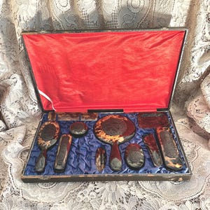 May include: A vanity set featuring a handheld mirror, brushes, a comb, and small containers with a tortoiseshell pattern and dark brown carved floral details. The items are arranged in a rectangular case with a bright red lid lining and blue base.