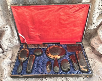 May include: A vanity set featuring a handheld mirror, brushes, a comb, and small containers with a tortoiseshell pattern and dark brown carved floral details. The items are arranged in a rectangular case with a bright red lid lining and blue base.