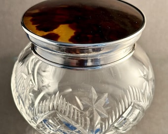 May include: A clear glass vanity jar featuring intricate star and cross-hatch cut patterns on the body. The jar is topped with a circular lid made of polished silver and a dark brown, mottled tortoiseshell-style inlay with a smooth finish.