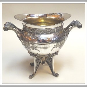 May include: Ornate silver-plated bowl with a wide, flared rim and two decorative handles. The bowl features intricate floral and vine patterns, and rests on three ornate legs. The interior has a golden hue.