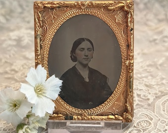 May include: An antique daguerreotype portrait of a person with dark hair styled back, wearing a dark garment with a small brooch, encased in an ornate, rectangular gold-toned metal frame with an oval center and intricate embossed floral patterns.