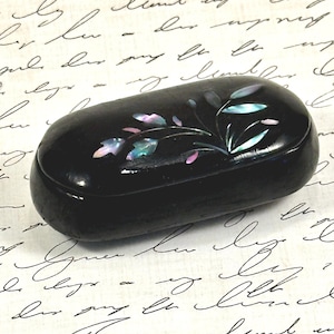 May include: An oval-shaped black lacquer box with a smooth, glossy finish. The lid features an inlaid mother-of-pearl design depicting a delicate floral sprig with iridescent leaves and petals in shades of blue, pink, and white.