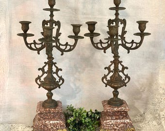 May include: A pair of five-arm candelabras featuring dark bronze-toned metal arms with ornate scrollwork and candle cups. Each candelabra rests on a square, mottled reddish-brown marble base supported by four decorative gold-toned metal claw feet.