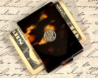 May include: A rectangular money clip featuring a dark brown and black tortoiseshell pattern. A circular silver-toned metal emblem is centered on the front surface, embossed with the letters GAP in a bold, capitalized, and sans-serif typeface.