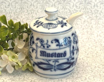 May include: A white ceramic mustard pot with a lid and a small spoon, featuring a blue floral pattern and the word Mustard printed on the front. The container has a rounded shape and a matching lid with a small knob handle on top.