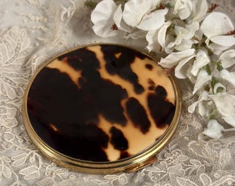 May include: A round compact mirror featuring a polished gold-tone metal frame and a smooth, glossy lid with a brown and cream tortoiseshell pattern. The circular case is closed and displays a high-contrast, mottled dark brown and beige design.