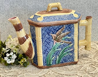 May include: A rectangular ceramic teapot featuring a blue basket-weave pattern, yellow bamboo-style trim, and a relief of a bird perched on green leaves. The spout has brown and yellow stripes, and the handle is shaped like a bamboo stalk.