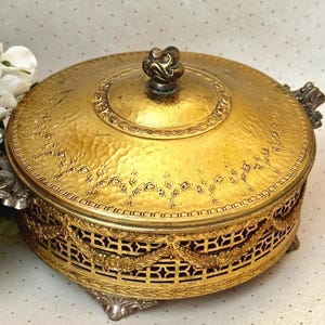 May include: A gold-colored, ornate jewelry box with a hammered texture on the lid. The box features intricate floral and geometric patterns, with decorative handles and feet. The lid is topped with a detailed knob.