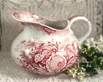 May include: A white ceramic pitcher featuring a detailed red transferware floral pattern. The vessel has a rounded body, a wide pouring spout, and a curved handle. The intricate botanical design covers the exterior surface with various flowers.
