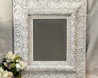 May include: Square, white, ornate mirror with a heavily carved floral design. The frame has a distressed, antique finish. A small bouquet of white flowers with green leaves is in the lower left corner.