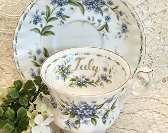 May include: A white porcelain teacup and saucer set featuring a delicate blue forget-me-not floral pattern and gold trim. The interior of the teacup displays the word July in elegant gold script, surrounded by a wreath of blue flowers and green leaves.