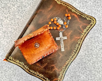 May include: A dark brown leather-bound book with gold embossed floral borders, featuring a small tan leather pouch with whip-stitched edges and a metal snap closure, accompanied by a rosary with orange beads and a silver-toned crucifix pendant.
