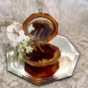 May include: An octagonal compact mirror with a brown and dark amber tortoiseshell pattern. The hinged case features a metal loop at the top and opens to reveal a reflective surface. The compact is displayed on a matching octagonal mirror base.
