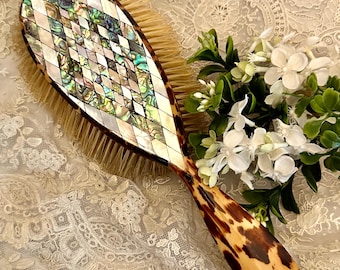 May include: A hairbrush featuring a handle and back with a brown and black tortoiseshell pattern. The back of the brush is inlaid with a diamond-shaped mosaic of iridescent abalone shell and white mother-of-pearl. The brush has light tan bristles.
