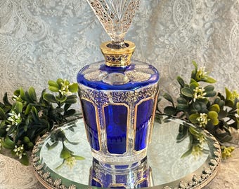 May include: A cobalt blue glass decanter featuring a clear glass stopper with a fan-shaped top. The decanter body has vertical panels with gold-colored filigree detailing and a gold-colored neck collar, resting on a circular mirrored pedestal base.