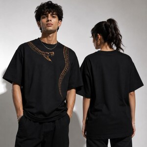 Puede incluir: Una camiseta negra de manga corta con un estampado gráfico de una serpiente enroscada en tonos marrones y dorados. El diseño de la serpiente comienza cerca del cuello y se extiende por el lado izquierdo del pecho. La parte trasera es negra lisa.