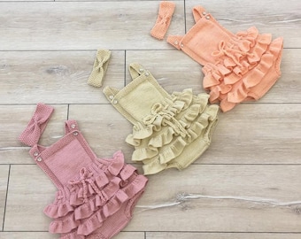 May include: Three knitted baby rompers in pink, peach, and beige, each with matching headbands. The rompers feature ruffled detailing and decorative ties at the chest. The outfits are displayed on a light wood-grain surface.