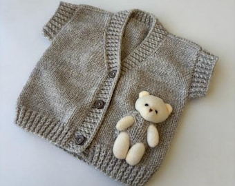 May include: A light gray knitted baby cardigan with short sleeves and three buttons down the front. A small, white teddy bear applique is sewn on the front. The cardigan is made of soft yarn.