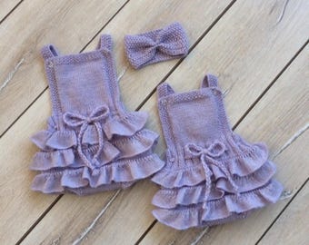 May include: Two lavender knitted baby rompers with ruffled bottoms and a matching headband with a bow. The rompers have straps and a bow at the waist. The set is displayed on a light wood surface.
