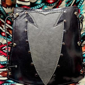 May include: Black leather shoulder bag with a gray, pointed design on the front. The bag is adorned with silver rivets and hardware. The bag has a black strap and is set against a colorful patterned fabric.