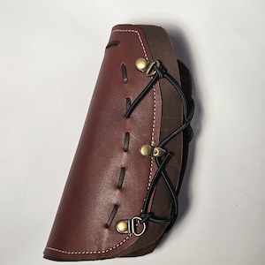 May include: A two-tone layered leather arm guard featuring a smooth reddish-brown outer layer with white stitching and a darker brown inner layer. It is secured with black cord lacing threaded through three antique brass-colored metal eyelets.