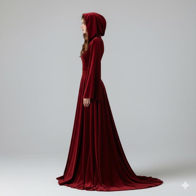 May include: A long, flowing, burgundy velvet gown with a hood. The dress features long, bell sleeves and a fitted waist. The skirt flares out dramatically, creating a dramatic silhouette. The dress is floor-length, with a slight train.