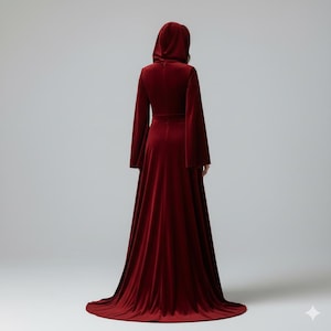 May include: A long, burgundy velvet hooded gown with flared sleeves and a floor-length skirt. The dress features a fitted bodice and a flowing train. The model is viewed from the back, showcasing the elegant design and rich color of the garment.