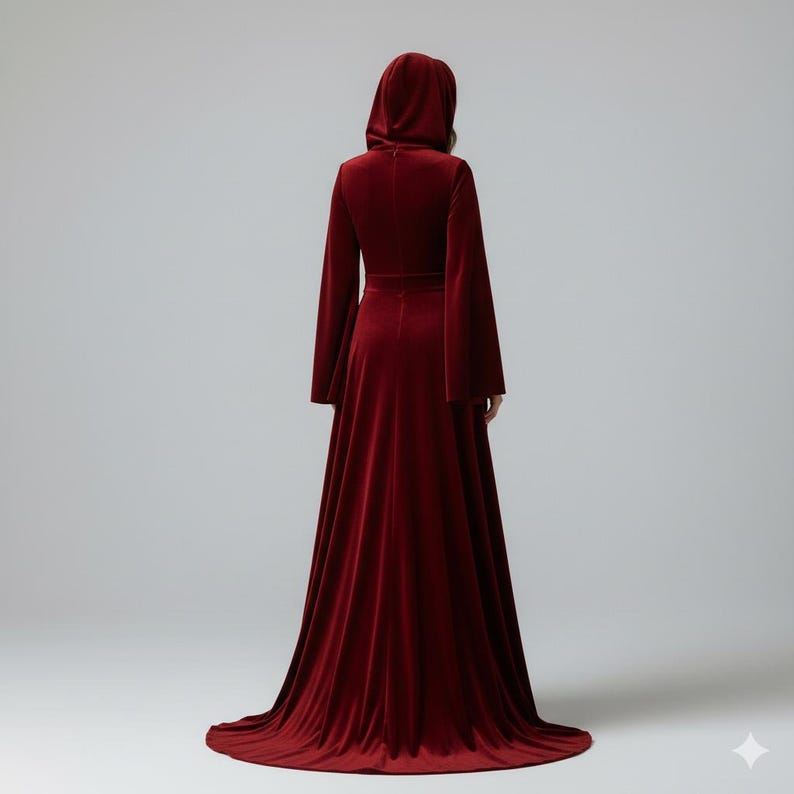 May include: A long, burgundy velvet hooded gown with flared sleeves and a floor-length skirt. The dress features a fitted bodice and a flowing train. The model is viewed from the back, showcasing the elegant design and rich color of the garment.