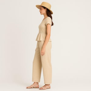 May include: A beige linen outfit comprising a short-sleeved top with a peplum, wide-leg trousers, a straw hat, and sandals. The outfit is a neutral colour and appears to be made of a lightweight fabric, suitable for warm weather.