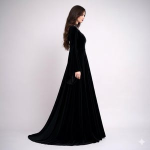 Pode incluir: Um vestido longo de veludo preto com uma saia fluida e mangas compridas e largas. O vestido tem um decote alto e uma cauda subtil. A modelo est&aacute; em perfil contra um fundo branco liso, mostrando a elegante silhueta do vestido.